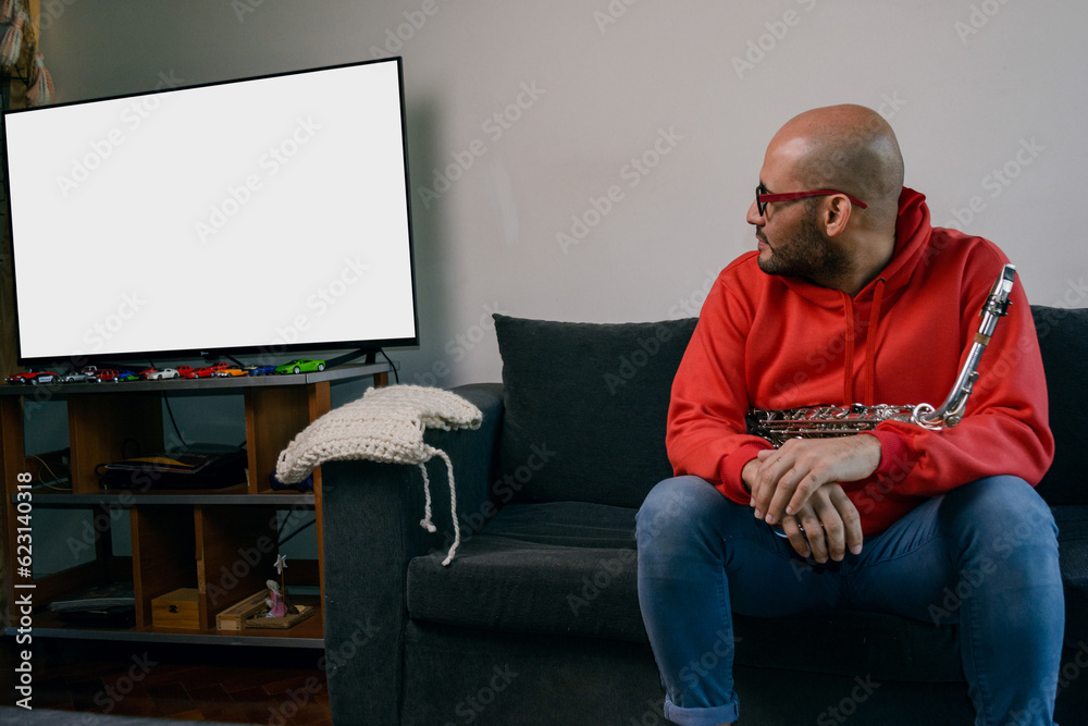 young latino musician man sitting with his saxophone at home turned to watch the tv screen. young latino musician man sitting with his saxophone at home turned to watch the tv screen.