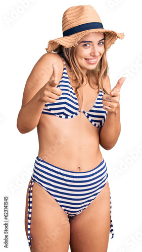 Young beautiful blonde woman wearing bikini and hat pointing fingers to camera with happy and funny face. good energy and vibes.