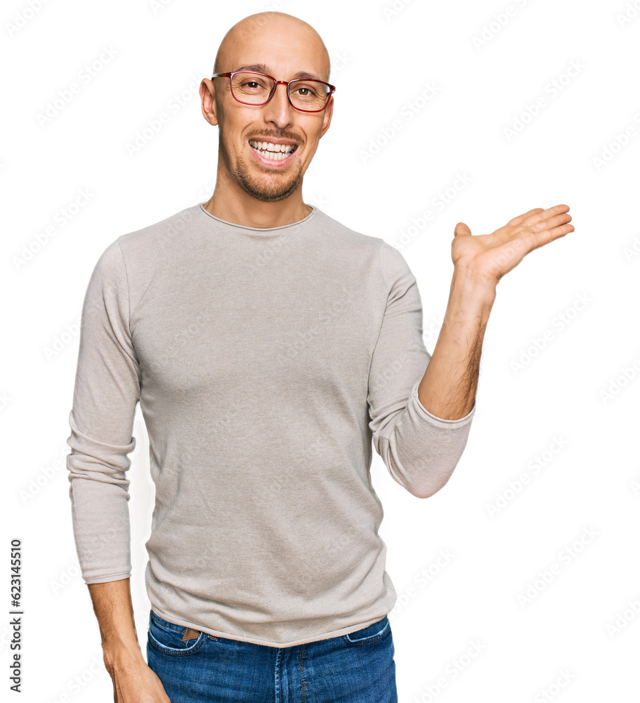 Bald man with beard wearing casual clothes and glasses smiling cheerful presenting and pointing with palm of hand looking at the camera.