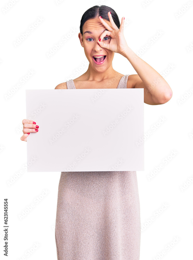 Beautiful brunette young woman holding blank empty banner smiling happy doing ok sign with hand on eye looking through fingers Beautiful brunette young woman holding blank empty banner smiling happy doing ok sign with hand on eye looking through fingers