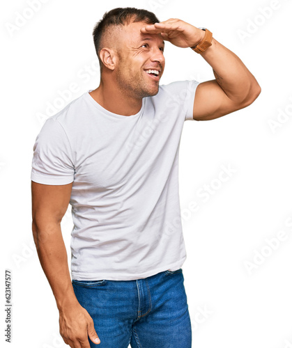 Handsome muscle man wearing casual white tshirt very happy and smiling looking far away with hand over head. searching concept.