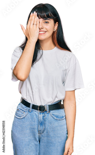 Young hispanic girl wearing casual clothes covering one eye with hand, confident smile on face and surprise emotion.