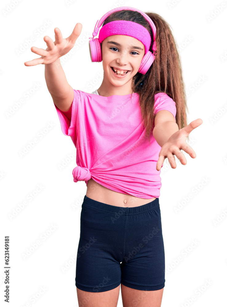 Cute hispanic child girl wearing gym clothes and using headphones looking at the camera smiling with open arms for hug. cheerful expression embracing happiness. Cute hispanic child girl wearing gym clothes and using headphones looking at the camera smiling with open arms for hug. cheerful expression embracing happiness.