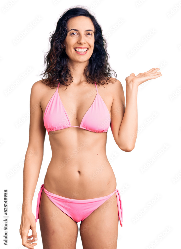 Young beautiful hispanic woman wearing bikini smiling cheerful presenting and pointing with palm of hand looking at the camera. Young beautiful hispanic woman wearing bikini smiling cheerful presenting and pointing with palm of hand looking at the camera.