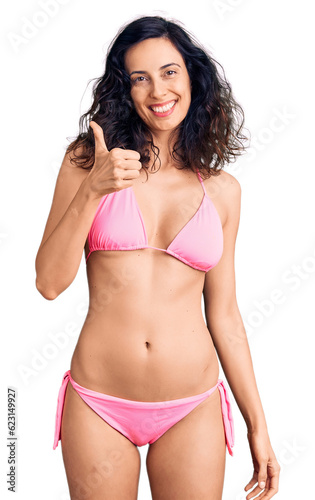 Young beautiful hispanic woman wearing bikini doing happy thumbs up gesture with hand. approving expression looking at the camera showing success. © Krakenimages.com