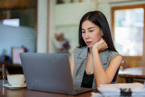 Owner of cafe is a beautiful Asian woman with long hair. Sit and work from boredom, put hands on chin at a table in a coffee shop. Use a laptop computer to examine the documents lying on the table.