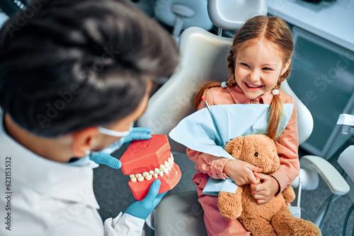фотографія A little girl sits in a dental chair, holds a toy rabbit in her hands, laughs and looks at the dentist who shows her a model of the jaw with teeth