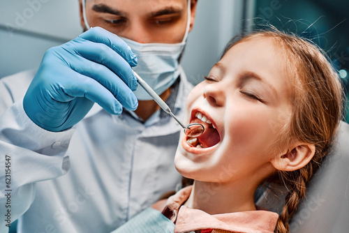 Checking teeth at a children's dentist. Close-up image of a doctor examining her teeth with a mirror, a small female patient sitting in a chair with her eyes closed.