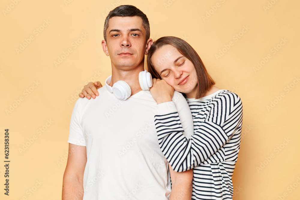 Smiling calm woman embracing serious man wearing casual outfits standing isolated over beige background female closed eyes felling love, guy looking at camera. Smiling calm woman embracing serious man wearing casual outfits standing isolated over beige background female closed eyes felling love, guy looking at camera.