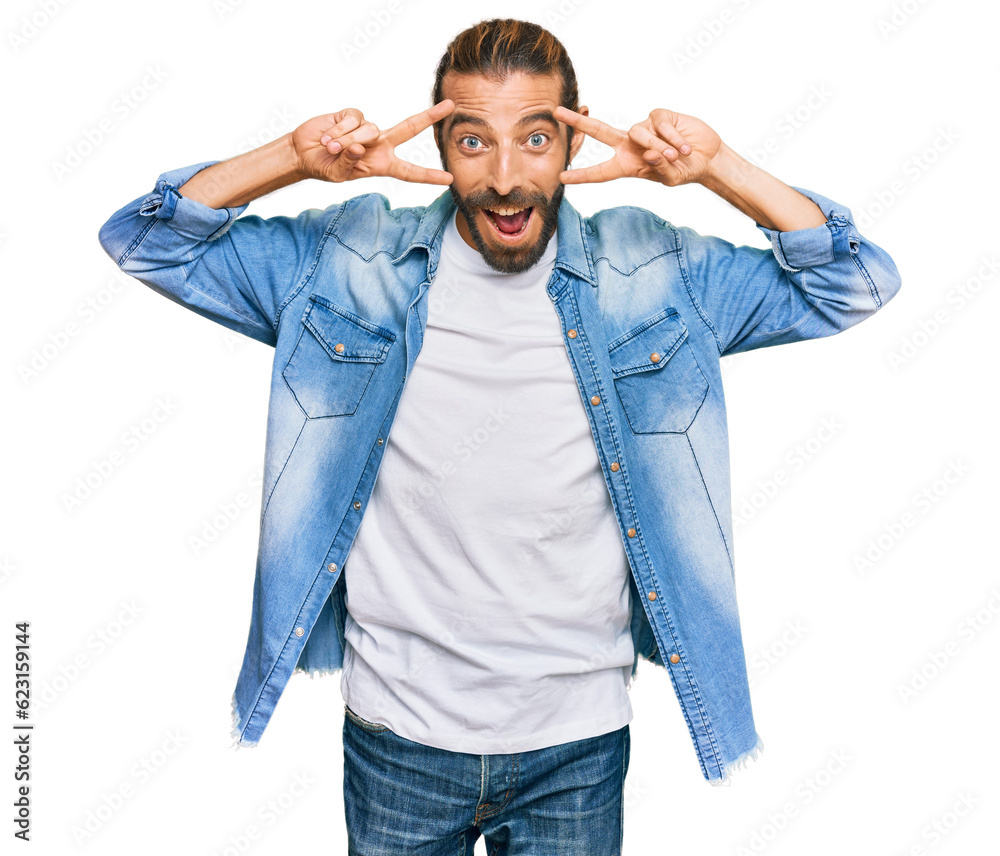 Attractive man with long hair and beard wearing casual denim jacket doing peace symbol with fingers over face, smiling cheerful showing victory Attractive man with long hair and beard wearing casual denim jacket doing peace symbol with fingers over face, smiling cheerful showing victory