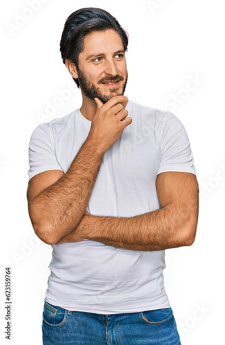 Young hispanic man wearing casual white t shirt with hand on chin thinking about question, pensive expression. smiling with thoughtful face. doubt concept.