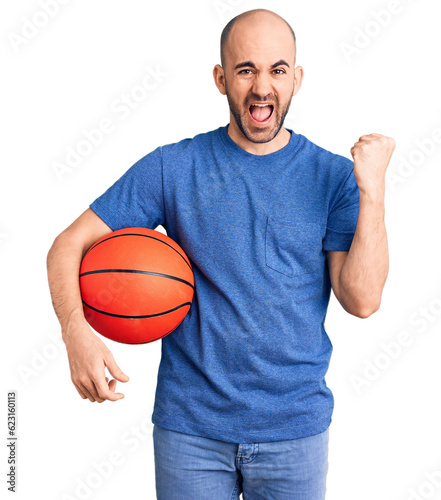 Young handsome man holding basketball ball screaming proud, celebrating victory and success very excited with raised arms