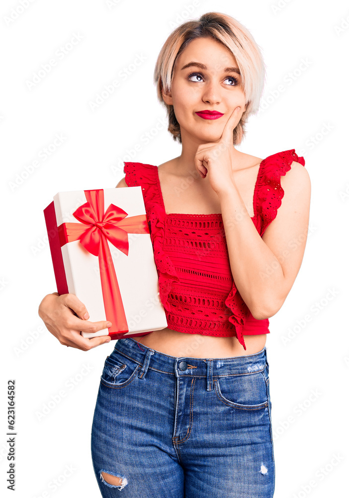 Young beautiful blonde woman holding gift serious face thinking about question with hand on chin, thoughtful about confusing idea Young beautiful blonde woman holding gift serious face thinking about question with hand on chin, thoughtful about confusing idea