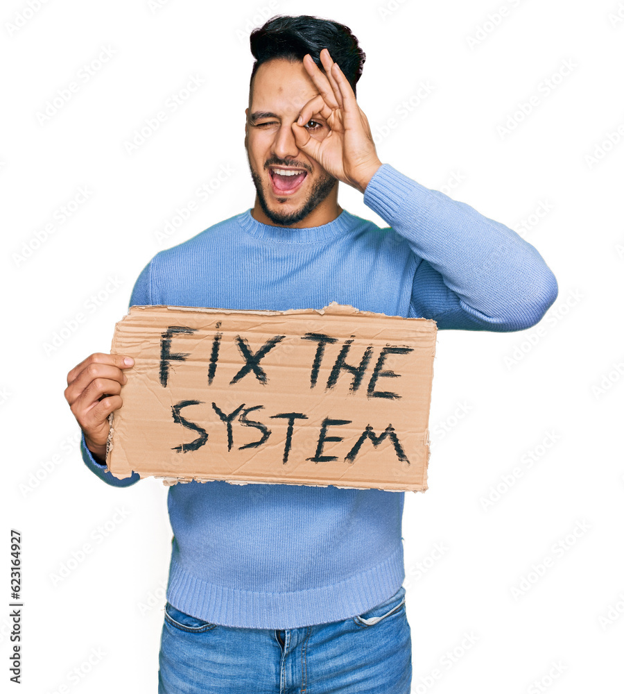 Young arab man holding fix the system banner cardboard smiling happy doing ok sign with hand on eye looking through fingers Young arab man holding fix the system banner cardboard smiling happy doing ok sign with hand on eye looking through fingers