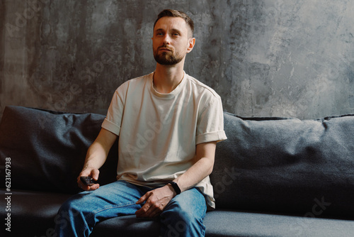 A handsome man with a beard in a white T-shirt and jeans holds a remote control from the TV and switches channels