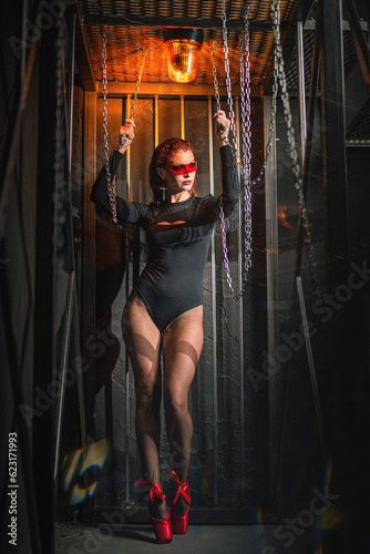 Beautiful young girl in the black body suit, stockings and red pointe shoes posing  on the metal constructions background in the yellow siren lights. Ballet dancer concept.
