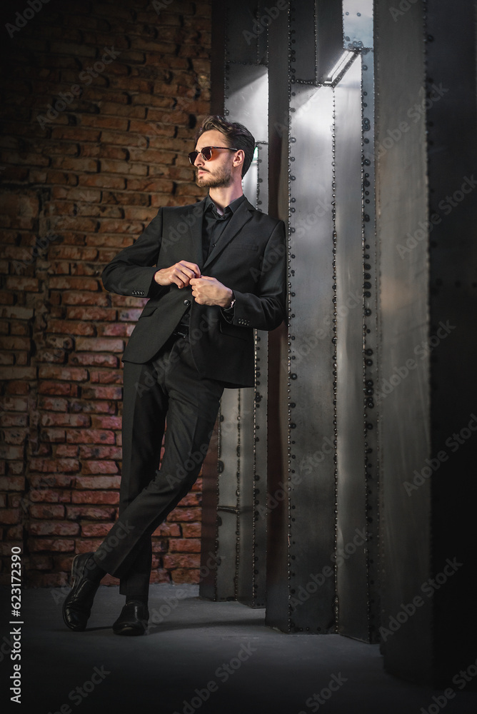 Serious man in classic black suit and sunglasses stand on the iron wall background.