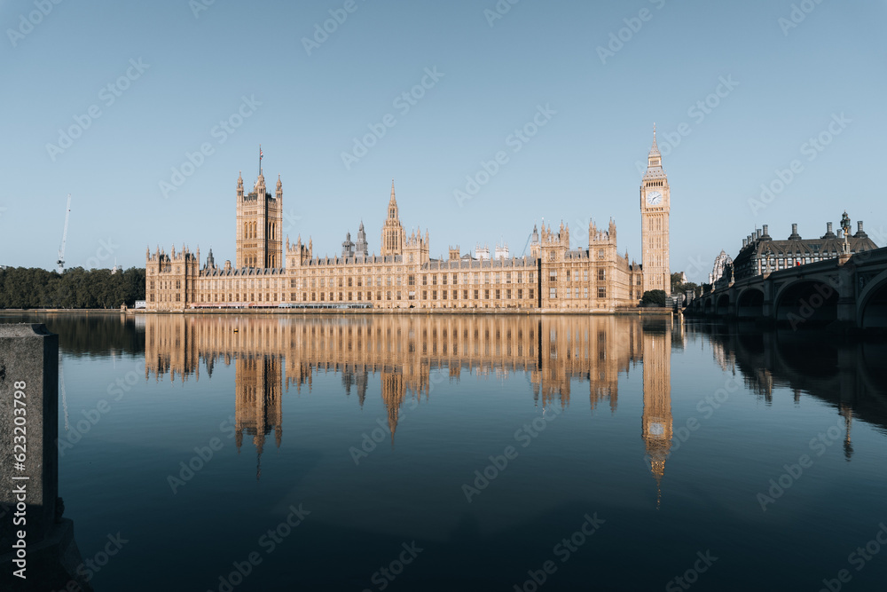 Photo of the entire English Parliament next to Big Ben. Photo taken from the other side of the river, with the reflection of the entire parliament in the river water. On a sunny day.