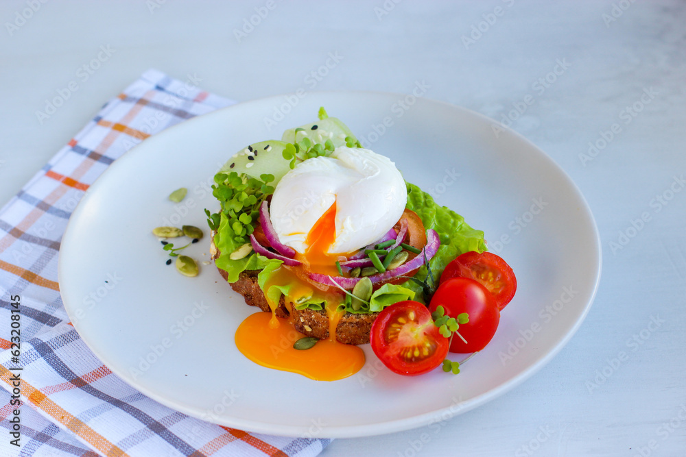 Runny poached egg on wholemeal bread with salad, tomato, red onion, lettuce and cucumber. Decorated with cherry tomatoes, microgreens, sesame and pumpkin seeds on white plate on cotton chessboard Runny poached egg on wholemeal bread with salad, tomato, red onion, lettuce and cucumber. Decorated with cherry tomatoes, microgreens, sesame and pumpkin seeds on white plate on cotton chessboard