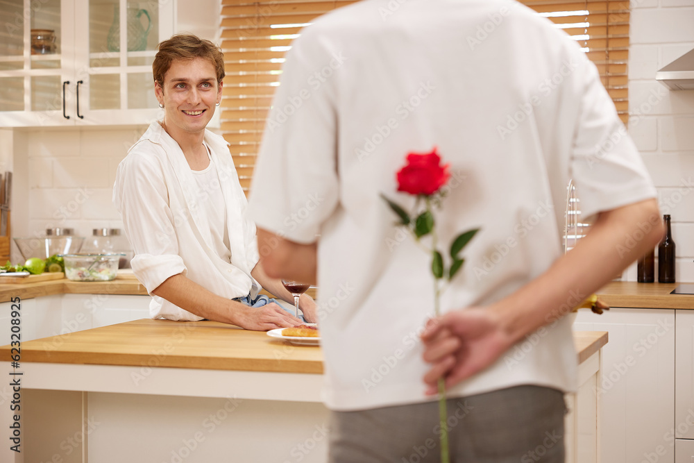 young gay couple looking at boyfriend and holding rose behind his back for surprise in the kitchen young gay couple looking at boyfriend and holding rose behind his back for surprise in the kitchen