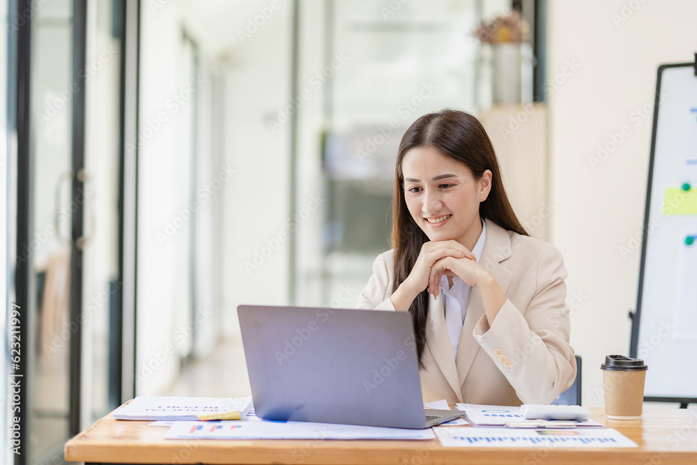 Asian female entrepreneur working in finance at home office analyzing financial graphs on documents with laptop on table in office