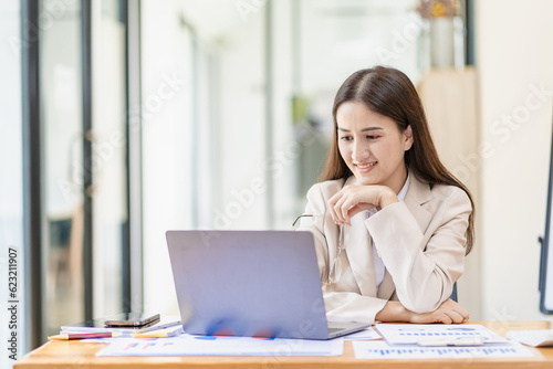 Asian female entrepreneur working in finance at home office analyzing financial graphs on documents with laptop on table in office