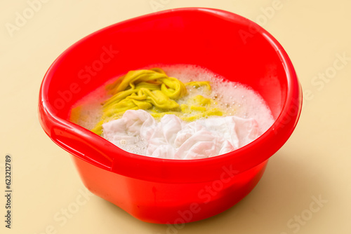 Plastic basin with dirty clothes, water and foam on yellow background, closeup