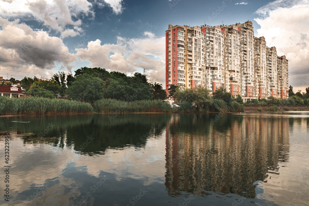 Reeds over the calm water of the lake. Summer city landscape. Cloudy sky over the lake. Picturesque view of the residential complex near the lake with reflection in the water. Reeds grow in the water.