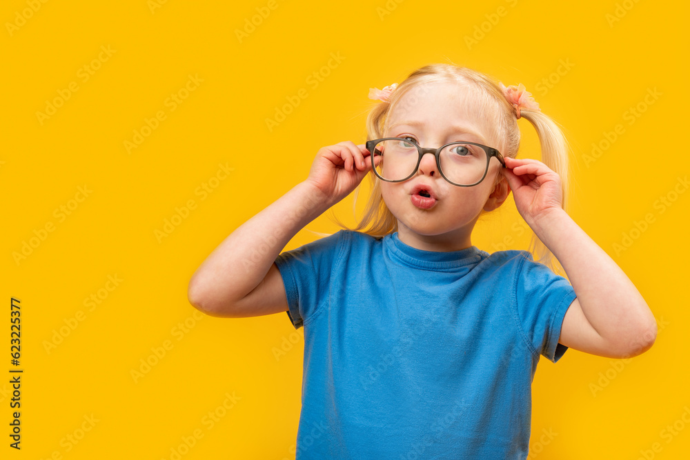 Little surprised girl adjusts her glasses. Portrait of blonde girl in blue T-shirt with glasses on yellow background.