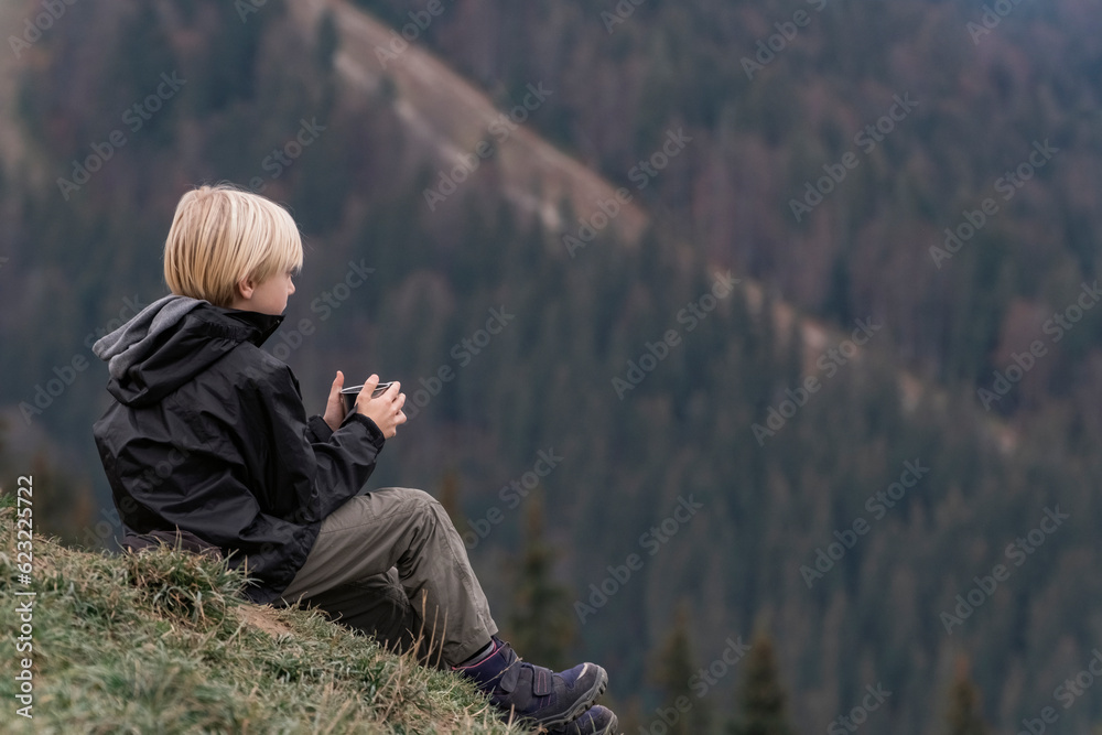 Portrait of boy in mountains resting on hillside with cup of tea. Hiking with child, tourist. Active recreation. Portrait of boy in mountains resting on hillside with cup of tea. Hiking with child, tourist. Active recreation.