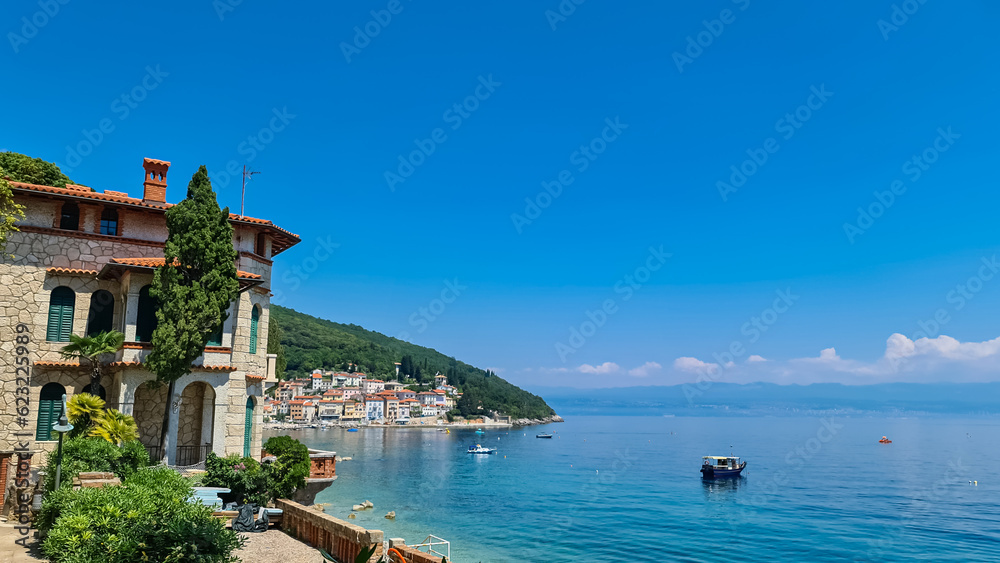 A panoramic view of the shore along Moscenicka Draga, Croatia. There is a small town located on the shore of the Mediterranean Sea. Few boats crossing the calm sea. Green hills in the back. Sunny day. A panoramic view of the shore along Moscenicka Draga, Croatia. There is a small town located on the shore of the Mediterranean Sea. Few boats crossing the calm sea. Green hills in the back. Sunny day.