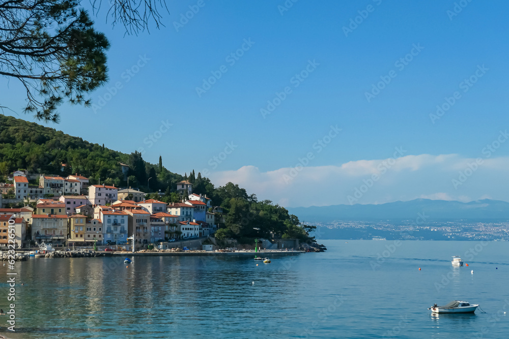A panoramic view of the shore along Moscenicka Draga, Croatia. There is a small town located on the shore of the Mediterranean Sea. Few boats crossing the calm sea. Green hills in the back. Sunny day.