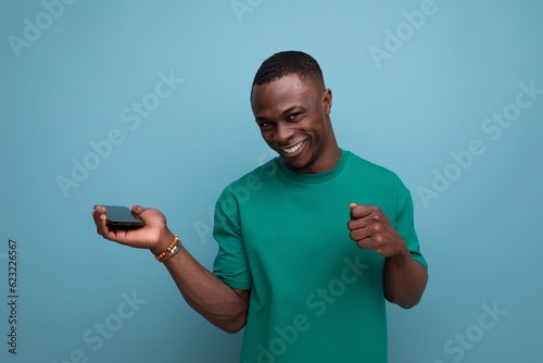 young smart african guy with short haircut dressed in a basic t-shirt holds a smartphone in his hand and smiles against the background with copy space