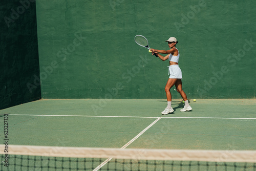 beautiful girl in a cap plays tennis on the tennis court against the background of a green wall © sutulastock