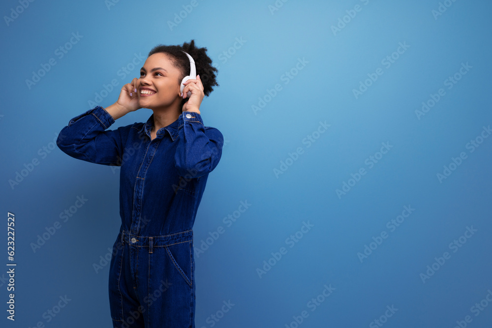 slim young hispanic brunette woman with fluffy curly hair in a blue denim suit with wireless headphones slim young hispanic brunette woman with fluffy curly hair in a blue denim suit with wireless headphones