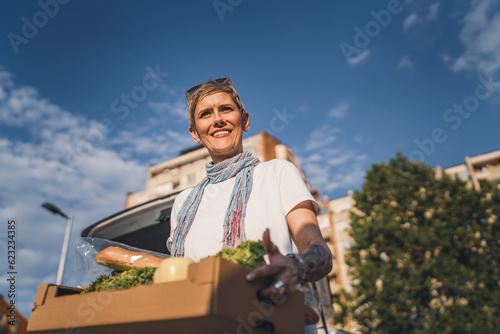 One woman mature caucasian female standing in the parking lot bu car in front of supermarket grocery store holding box with food and vegetables in sunny day happy smile healthy eating vegan concept © Miljan Živković
