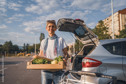 One woman mature caucasian female standing in the parking lot bu car in front of supermarket grocery store holding box with food and vegetables in sunny day happy smile healthy eating vegan concept