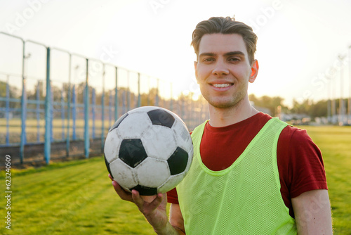 Man holding a soccer ball looking at the camera and smiling in the backlight of the sun. Portrait of man football soccer player at sunset. © glebcallfives