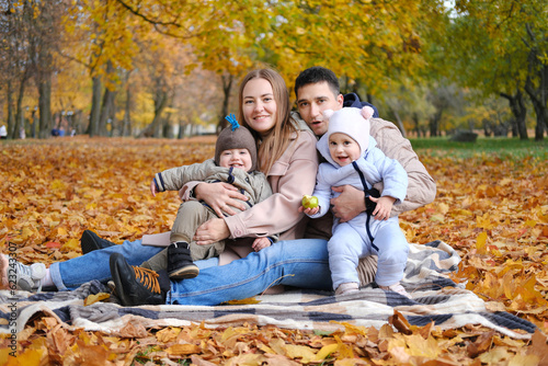 Parents and their little twin children are relaxing in the autumn park sitting on a blanket