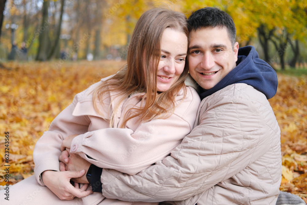 A young couple, husband and wife, sit on a blanket in an autumn park, the husband hugs his wife, smiles and looks at the camera A young couple, husband and wife, sit on a blanket in an autumn park, the husband hugs his wife, smiles and looks at the camera