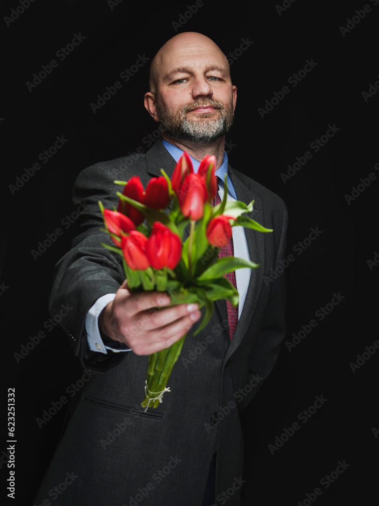 Man holding fresh red tulip bouquet in his hands. Model is bald with grey beard, wearing classic grey suit. Handsome male with special gift. Making up concept. Dark color background. Man holding fresh red tulip bouquet in his hands. Model is bald with grey beard, wearing classic grey suit. Handsome male with special gift. Making up concept. Dark color background.