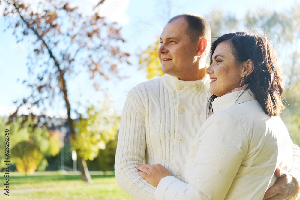 Husband and wife in white warm clothes pose for the camera in an autumn park on a sunny day. couple looking away Husband and wife in white warm clothes pose for the camera in an autumn park on a sunny day. couple looking away