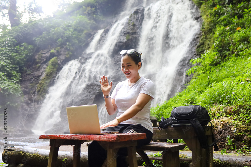 a female freelancer working in nature with a waterfall in the background.  remote working and online video working. 