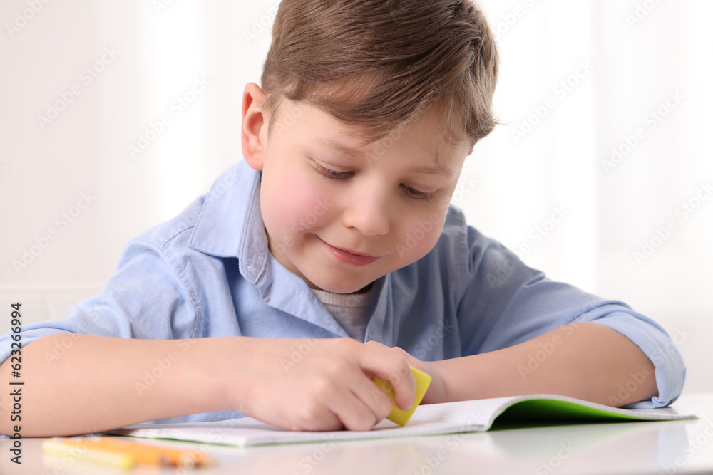 Little boy erasing mistake in his notebook at white desk indoors Little boy erasing mistake in his notebook at white desk indoors