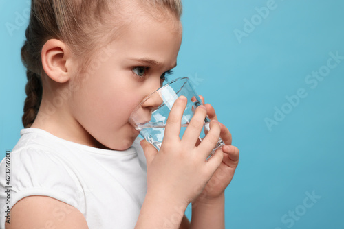 Cute little girl drinking fresh water from glass on light blue background, space for text