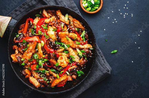 Asian cuisine stir fried chicken, paprika, mushrooms, chives with sesame seeds in frying pan. Black kitchen table background, top view