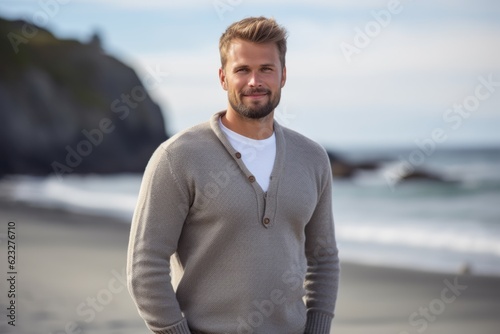 Portrait of handsome man standing on beach with hands in pockets smiling at camera