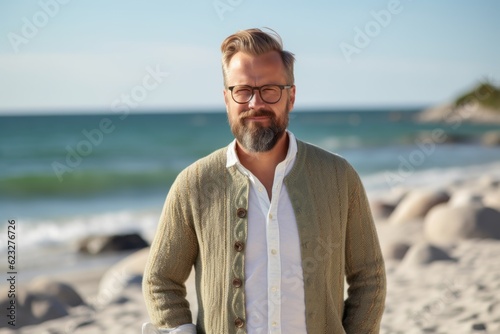 Handsome mature man with beard wearing casual clothes and glasses standing on the beach