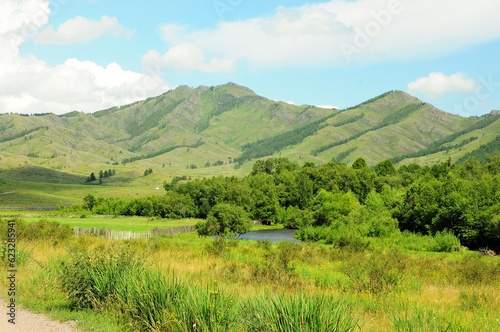 A wood-fenced pasture on the banks of a small meandering river with tall bushes on the banks and a range of hills in the background.