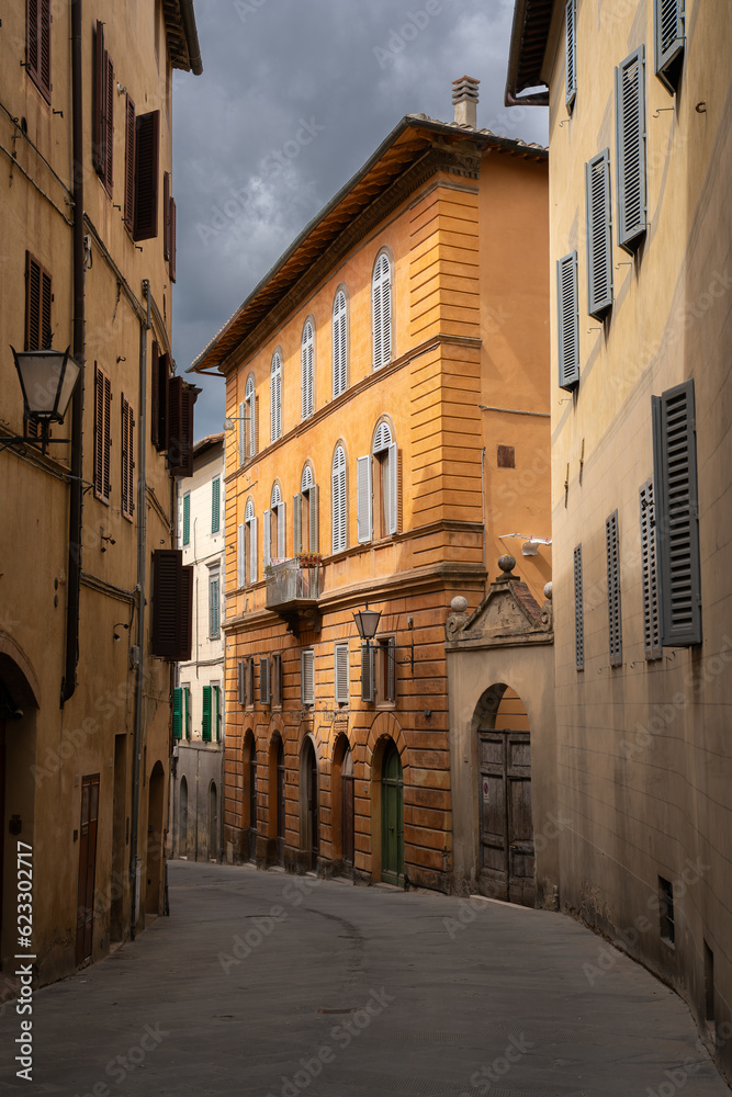 Moody Italian alleyway with sunlight illuminating a terracotta building and a gray sky in the background in the medieval city of Siena, Italy.